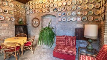 A cozy room with a stone wall, a dining table with chairs, a sofa with patterned cushions, and a wall decorated with numerous plates.