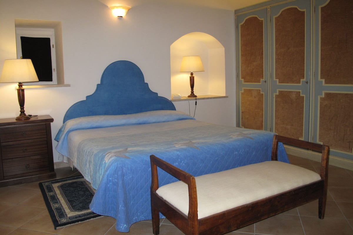 A bedroom with a bed, bedside lamps, a wooden chair, a nightstand, and a wall-mounted shelf.