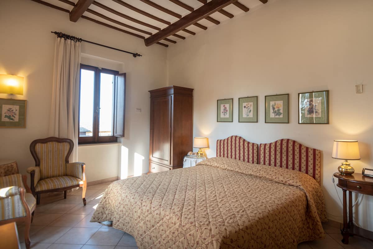 A bedroom with a large bed, a wooden wardrobe, a chair, a nightstand with a lamp, and framed pictures on the wall.