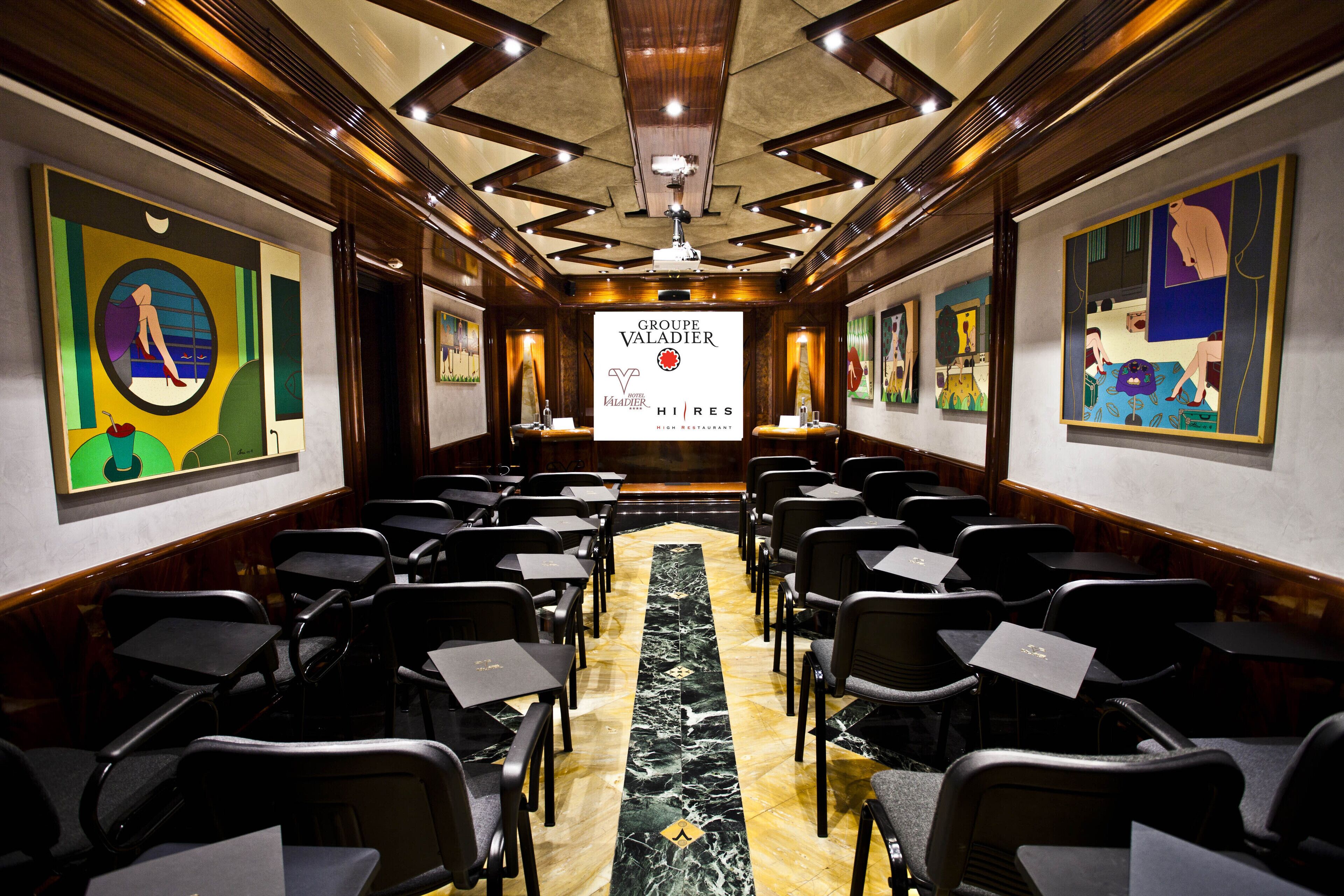 A conference room with rows of chairs, a central projection screen, and artwork on the walls.