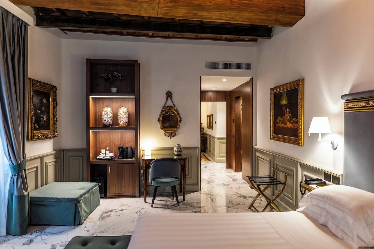 Une chambre d’hôtel avec un lit, un bureau, une chaise et une petite table avec une lampe. Les murs sont ornés de tableaux encadrés, et une étagère en bois présente divers objets décoratifs.