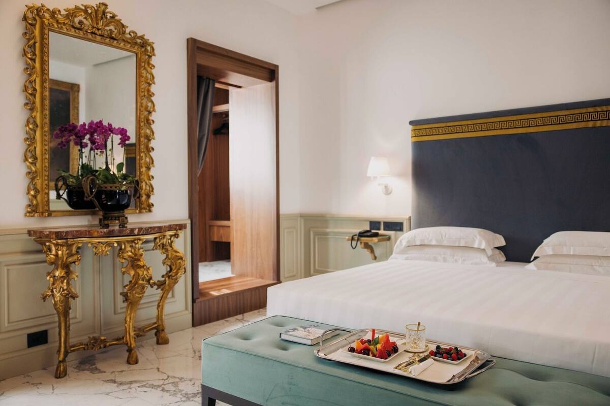 Une chambre d’hôtel avec un grand lit, une table de chevet sur laquelle se trouve une assiette de fruits, une coiffeuse avec un miroir et un vase de fleurs, et une porte donnant sur une autre pièce.