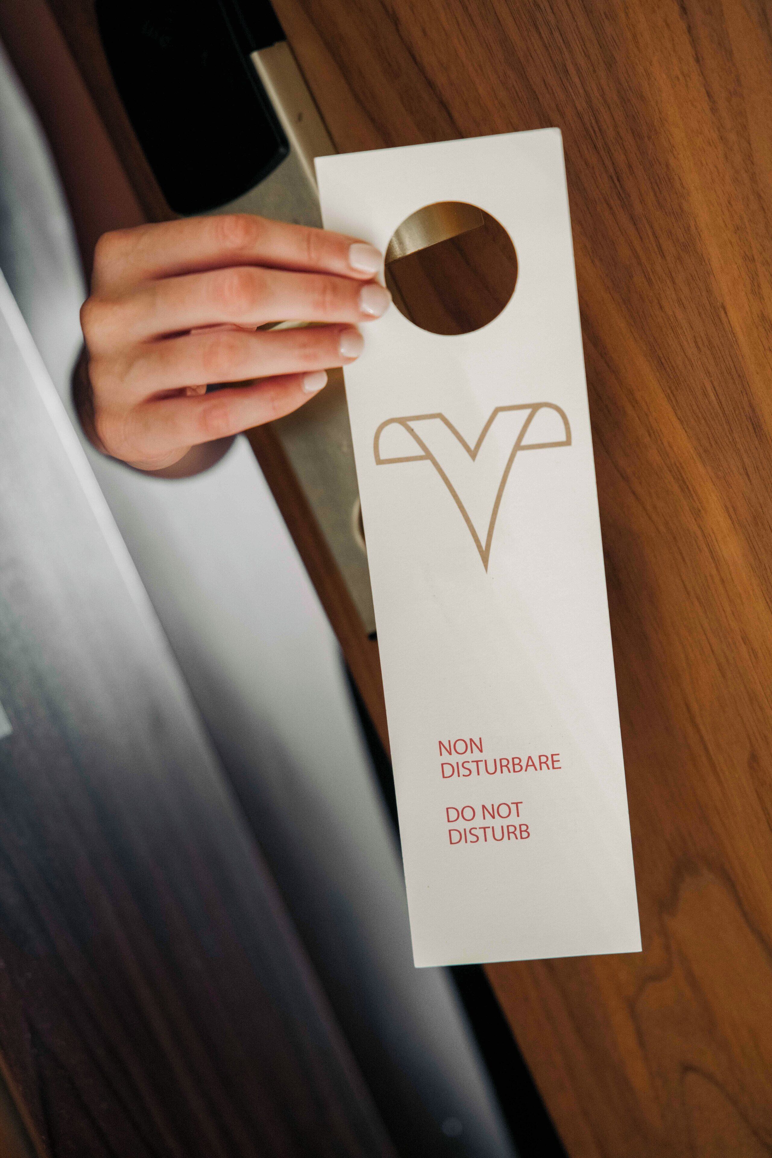 A hand holding a white box with a circular cutout and a logo, containing text that reads "NON DISTURBARE DO NOT DISTURB".