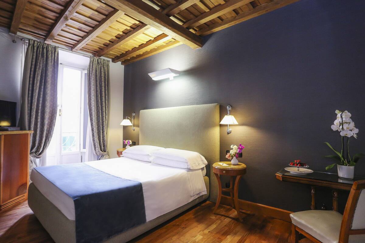 Une chambre d’hôtel avec un grand lit, des poutres apparentes en bois au plafond, deux lampes de chevet, une petite table sur laquelle est posé un vase de fleurs et un fauteuil.