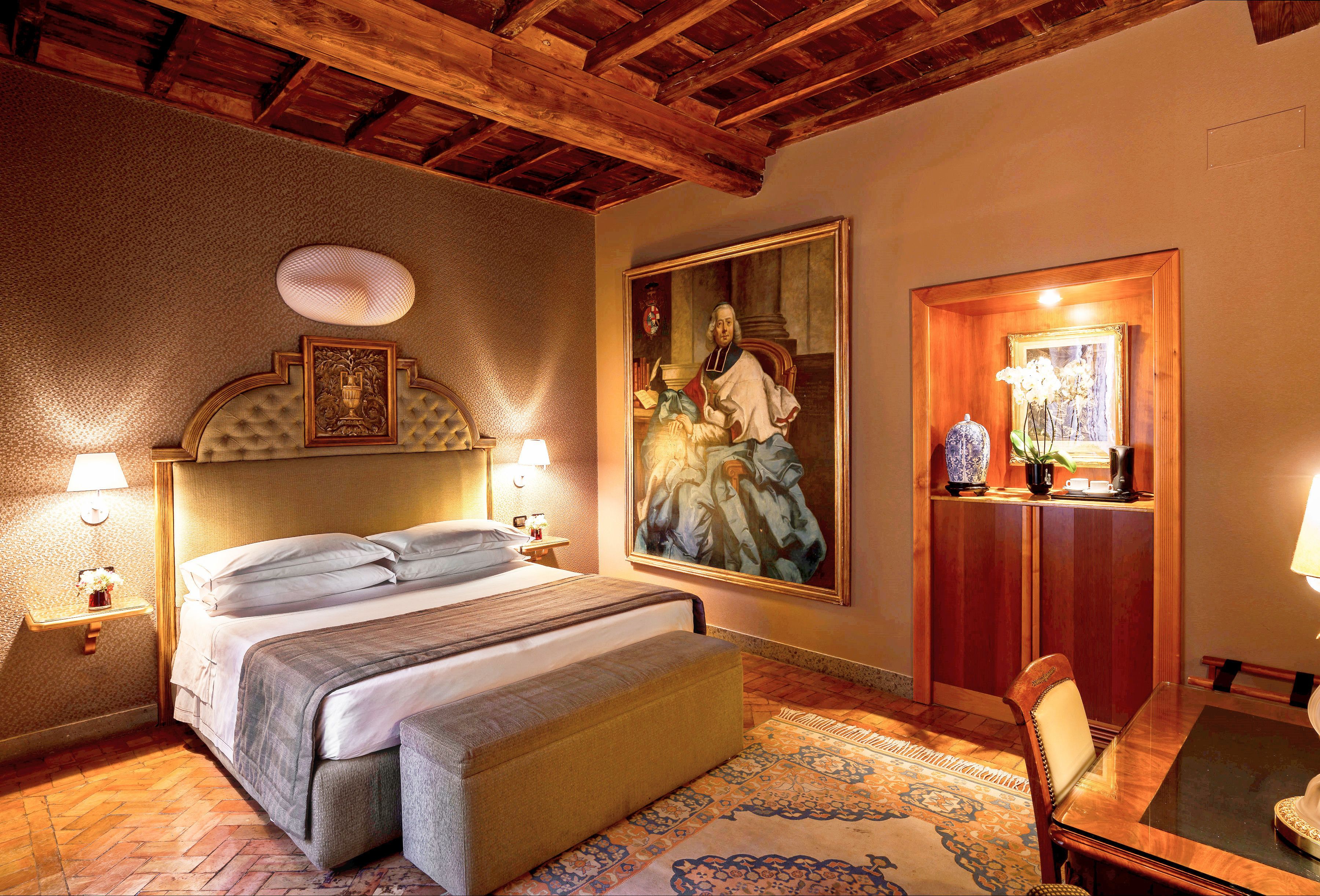 A hotel room with a large bed, a wooden ceiling, a painting on the wall, and a small table with a lamp.