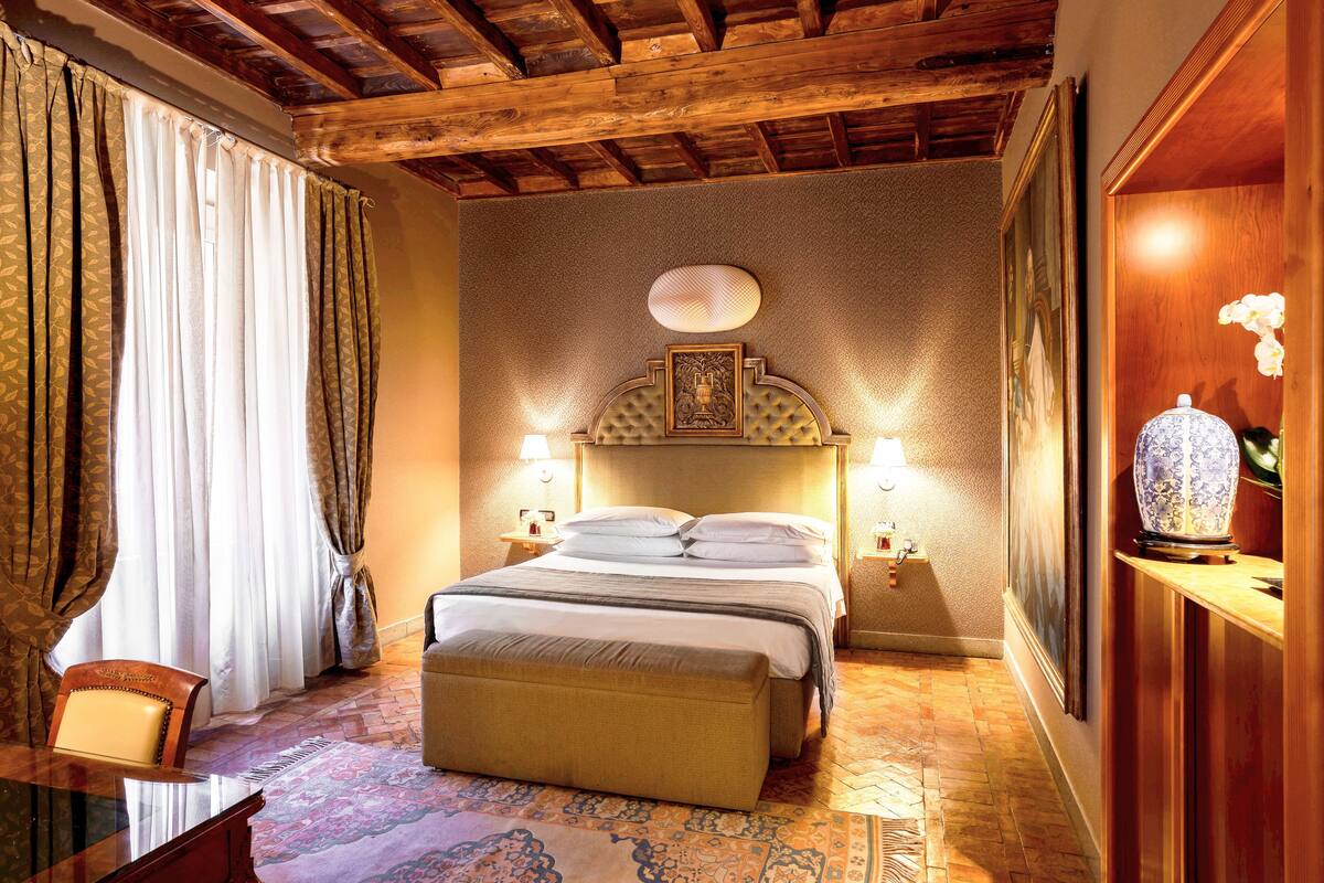 Une chambre d’hôtel avec un plafond en bois, un lit doté d’une tête de lit, deux lampes de chevet, une table de chevet, une chaise et un vase décoratif.