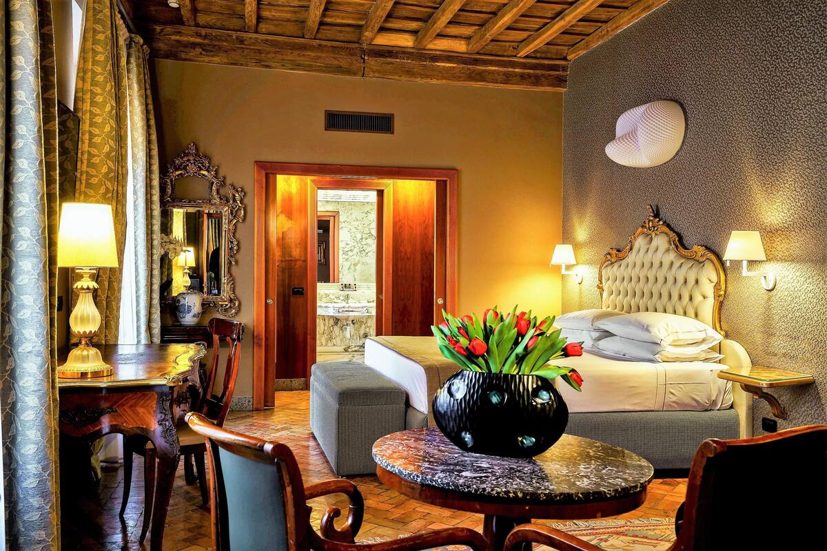Une chambre d’hôtel avec un lit, une table ronde ornée d’un vase de fleurs, une chaise, un bureau avec une lampe et un miroir.