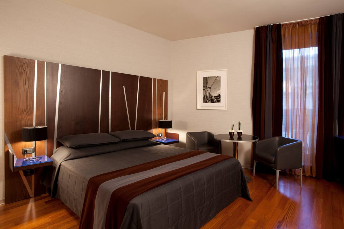 Une chambre d’hôtel moderne avec un grand lit, des tables de chevet avec des lampes, une petite table ronde, une chaise et une fenêtre avec des rideaux.