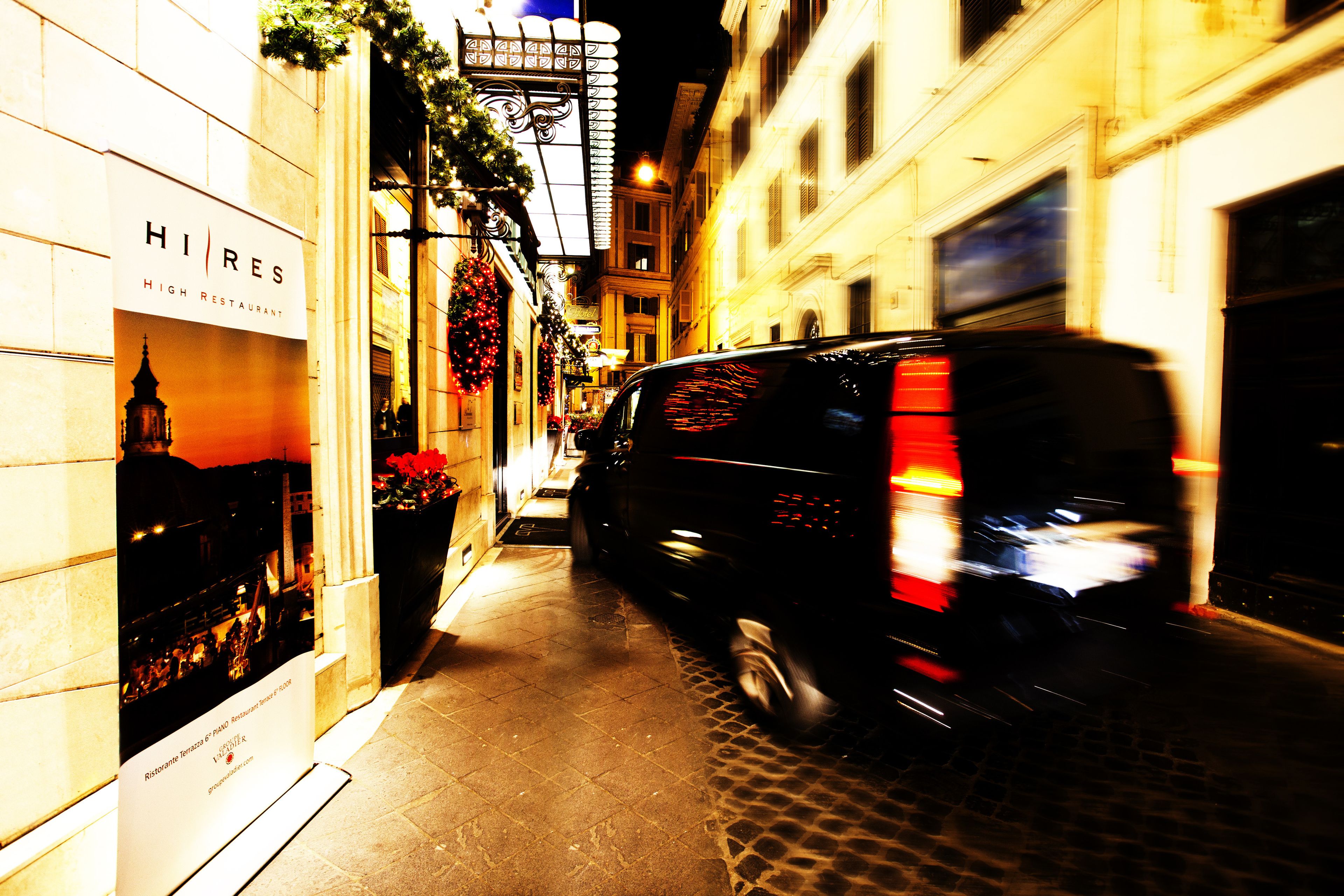 A narrow street with a black van parked on the right, a building with a 'HIRES' sign, and a Christmas decoration.