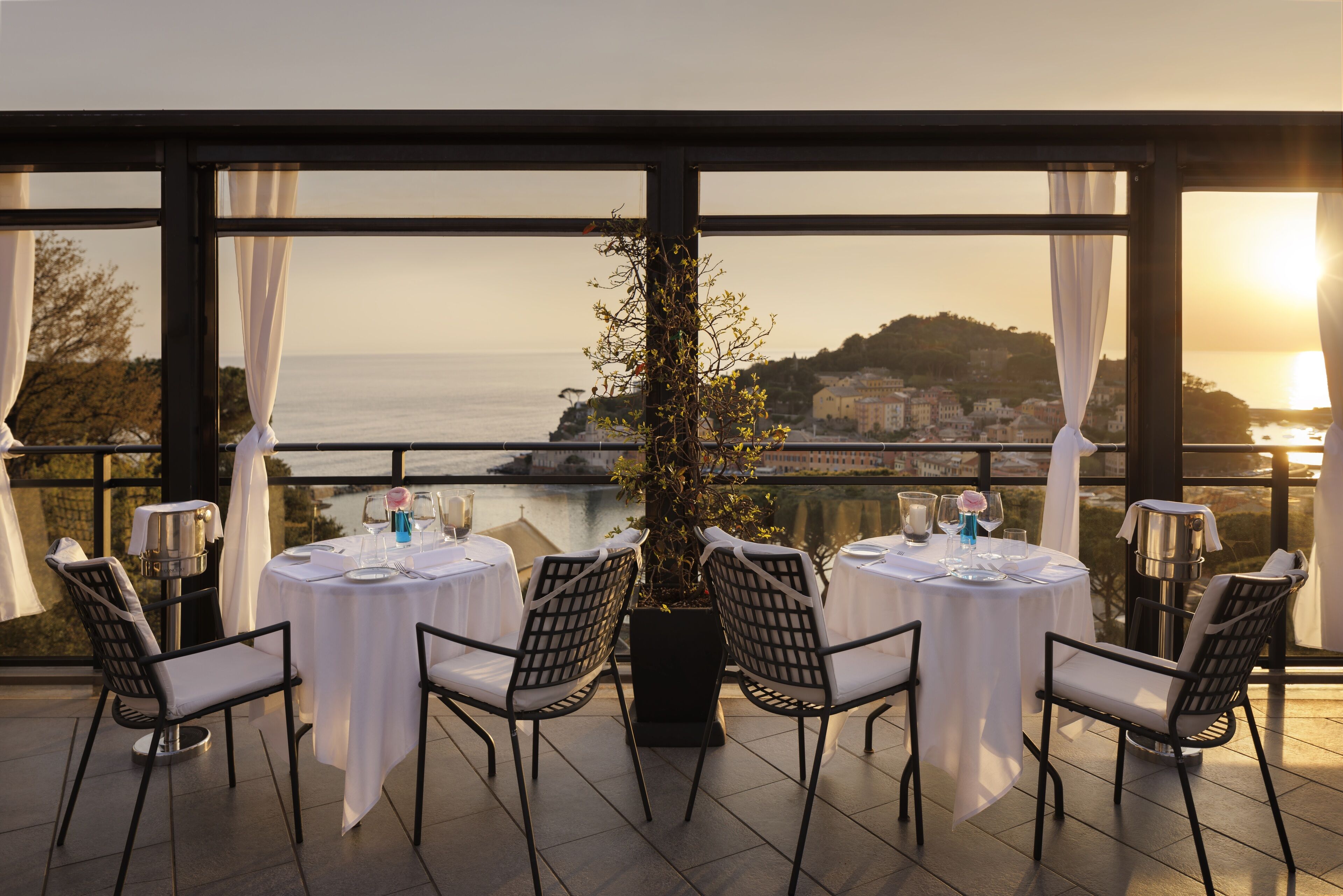 Terraço com quatro mesas preparadas para refeições, com vista para a água e para a paisagem urbana.