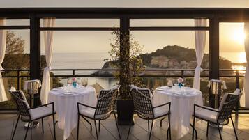 Terraço com quatro mesas preparadas para refeições, com vista para a água e para a paisagem urbana.