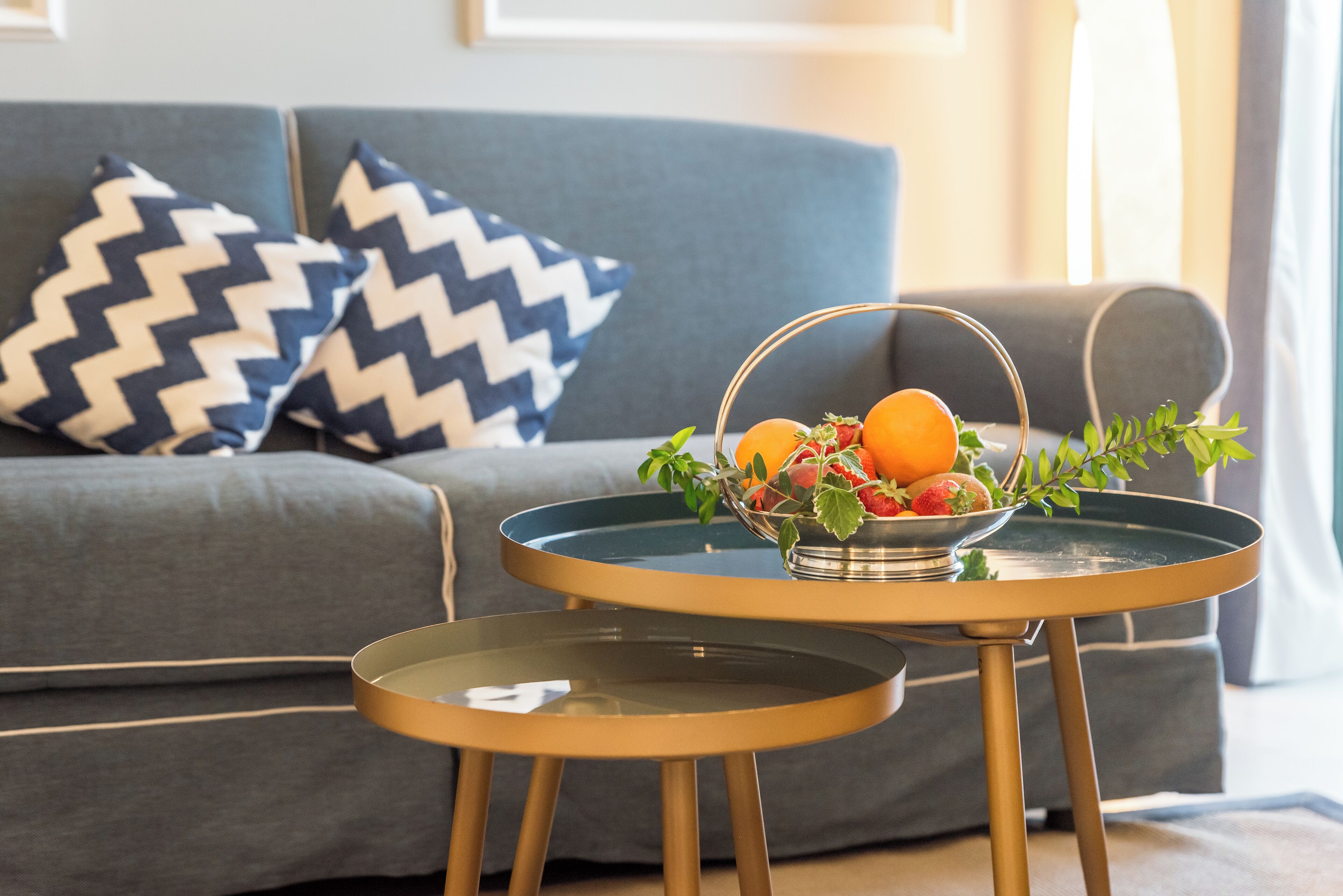 A modern living room with a grey sofa, two gold tables, and a fruit basket on one table.