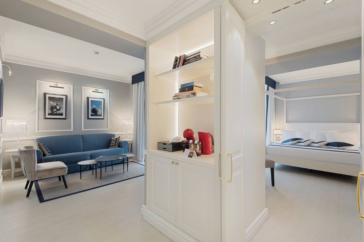 A modern living room with a blue sofa, a white cabinet with shelves, and a bed with white bedding.