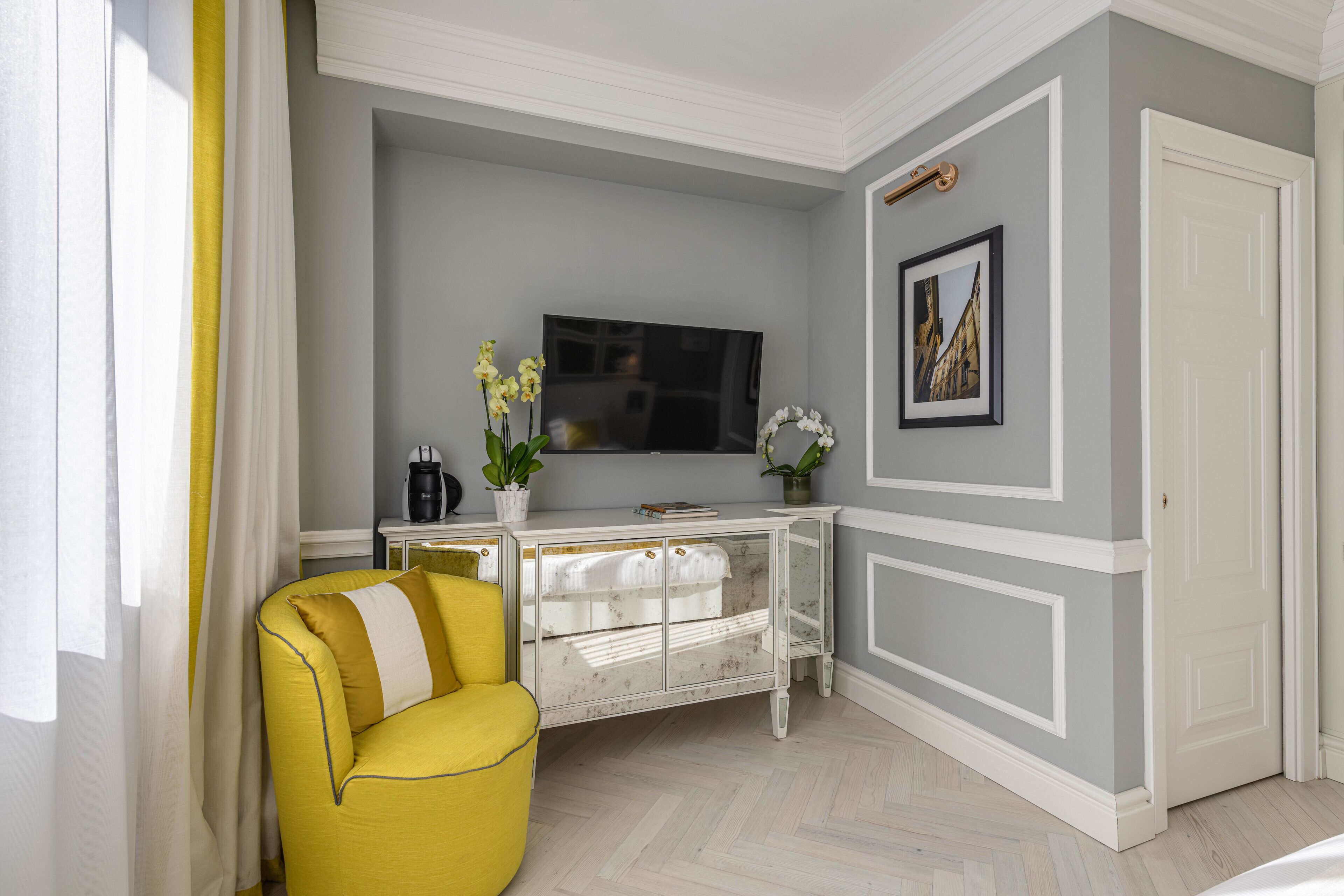 Una habitación con un sillón amarillo, un escritorio blanco con televisor de pantalla plana, una planta en maceta y un cuadro en la pared.