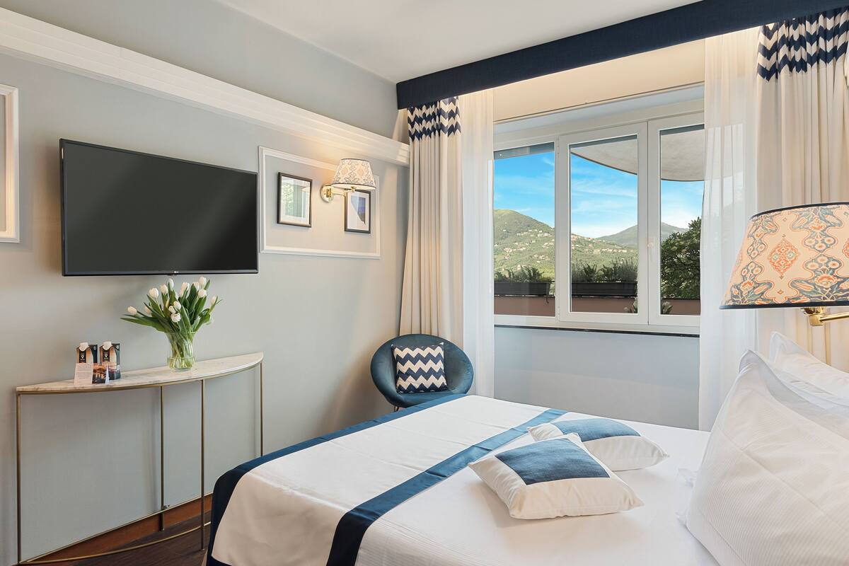A hotel room with a bed, a TV, a desk with a vase of flowers, and a view of the outdoors.