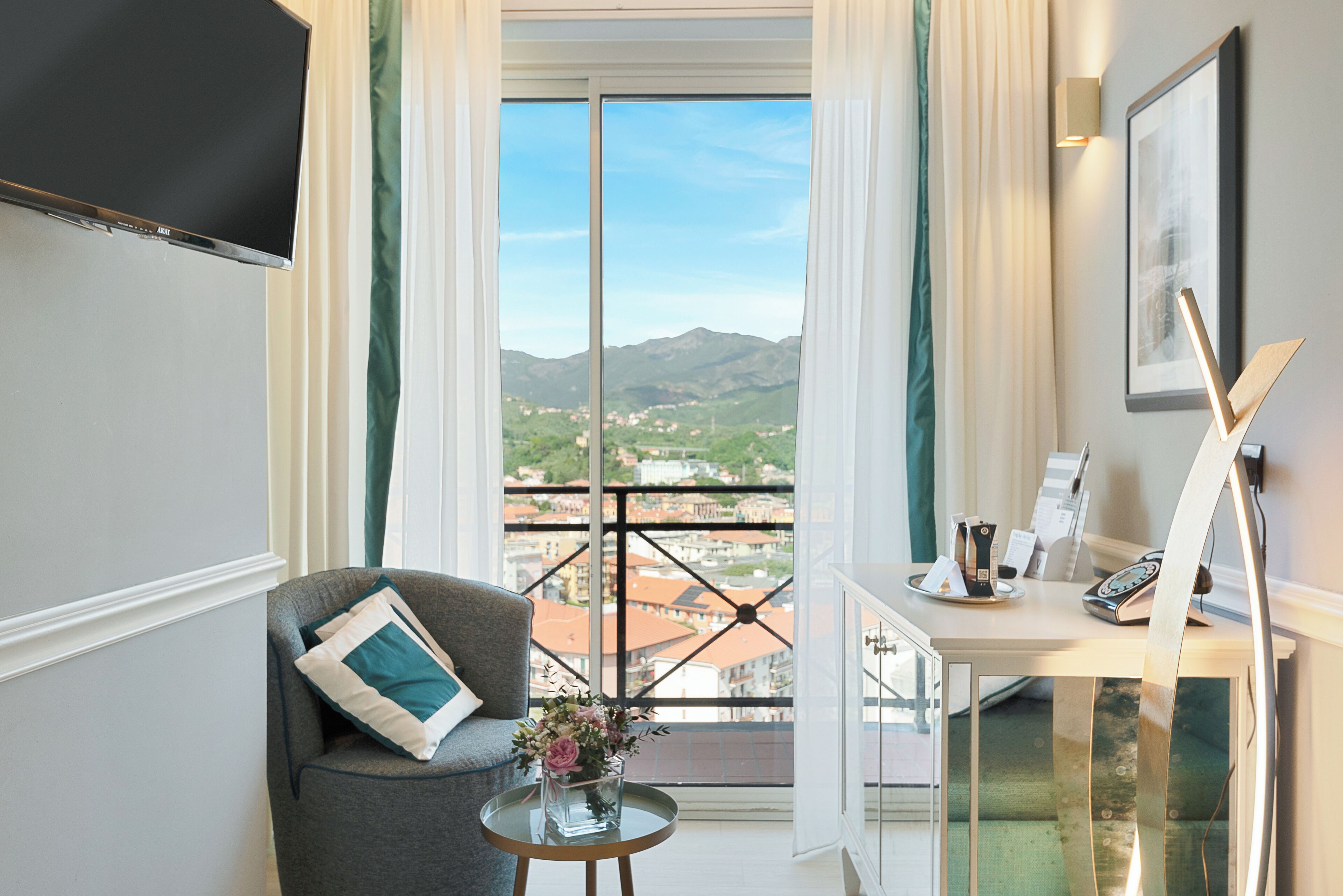 A room with a balcony, a chair, a small table with a vase of flowers, and a desk with a telephone and papers.