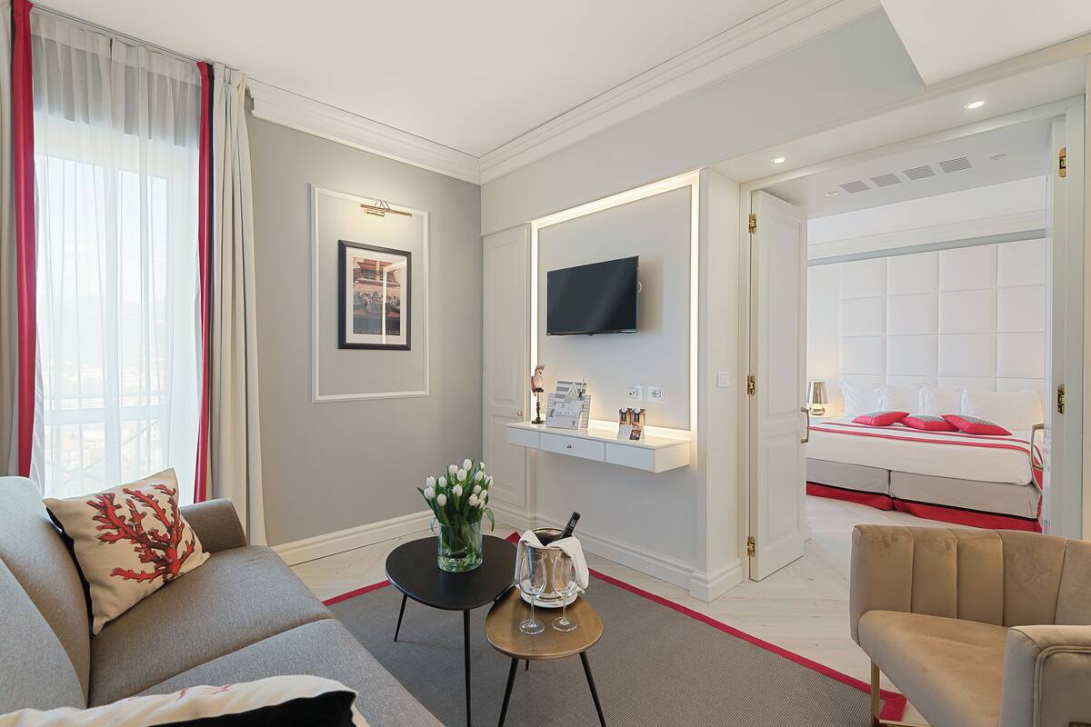 A modern hotel room with a sofa, a small table with a vase of flowers, a flat-screen TV mounted above a bedside table, and a bed with red bedding.