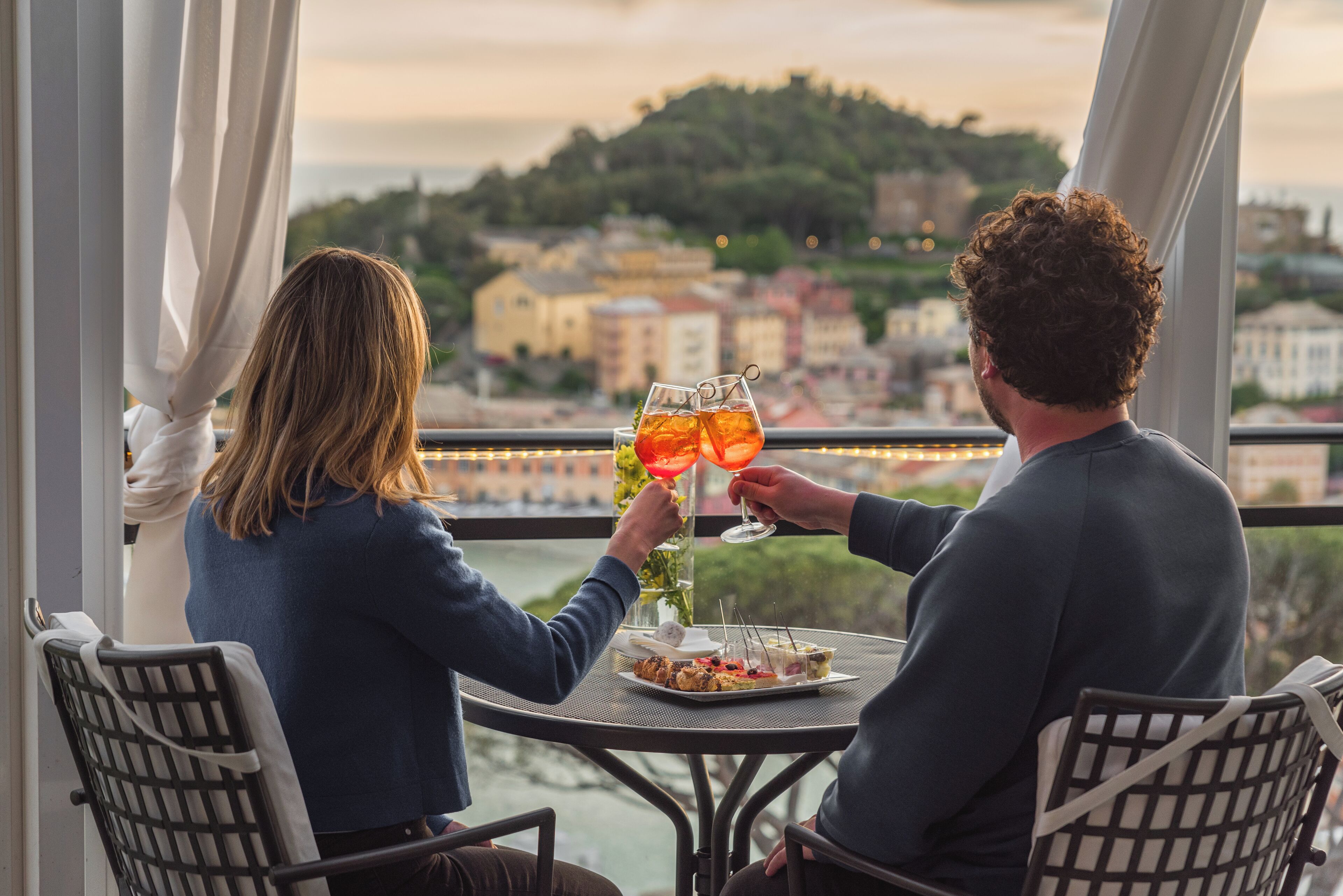 Two people seated at a table with a city view, holding glasses with drinks.