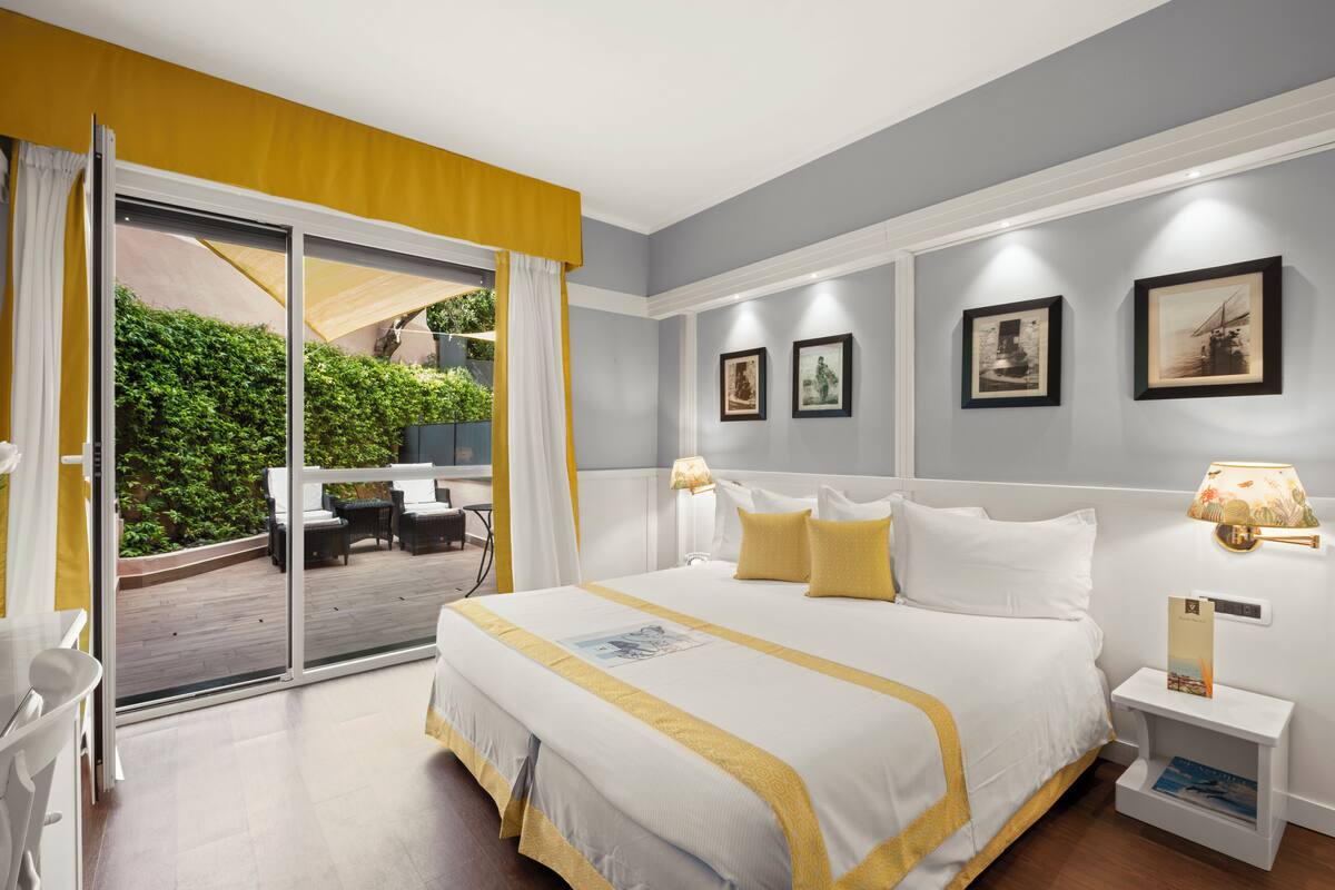 A hotel room with a bed, a desk, a chair, a small table, and a view of a garden through a sliding door.