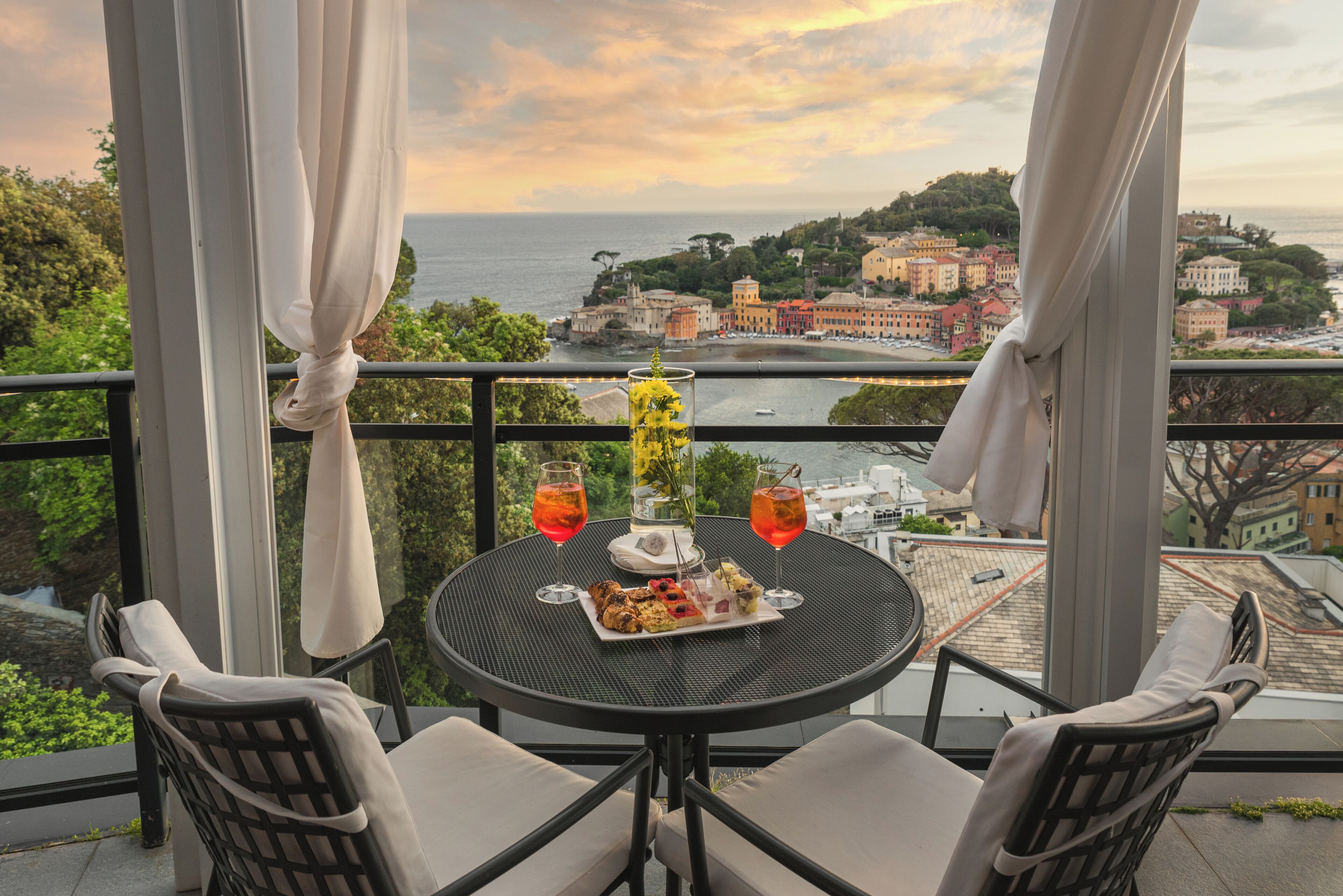 Un balcon avec une table dressée pour deux personnes, comprenant une assiette de nourriture et deux verres de vin rouge, donnant sur une ville côtière.