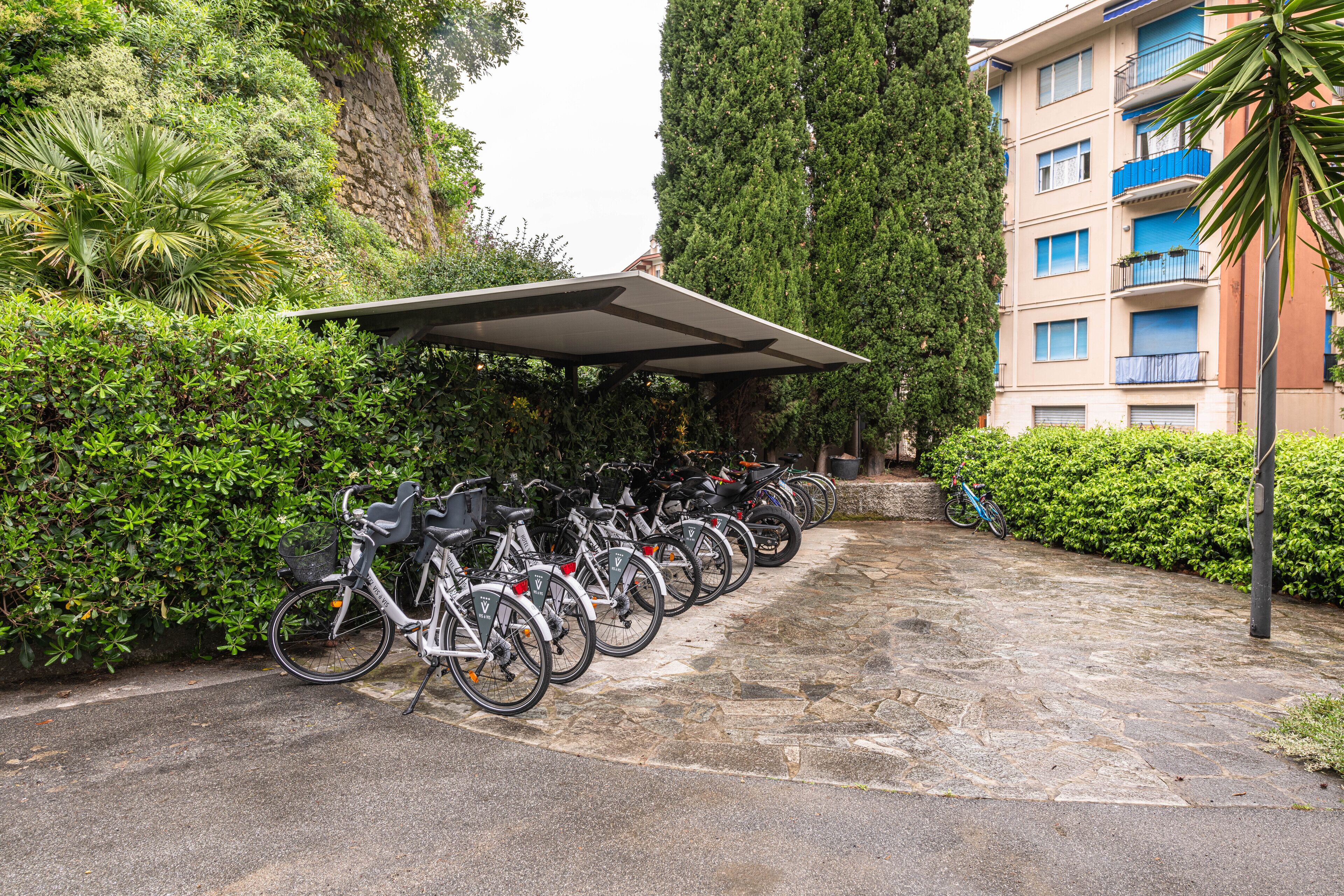 A row of bicycles parked under a covered area, surrounded by greenery and a building with balconies.