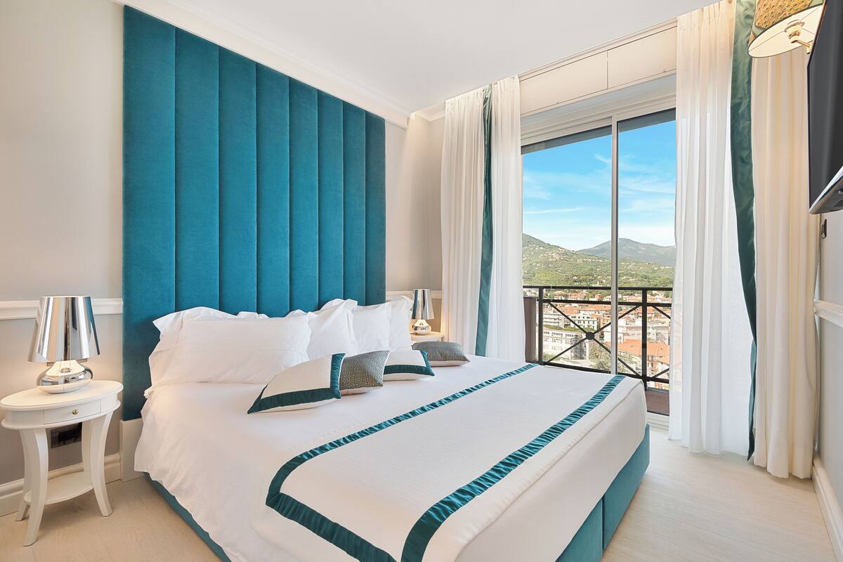 A hotel room with a large bed, a nightstand, a lamp, and a view of a mountainous landscape through a balcony.