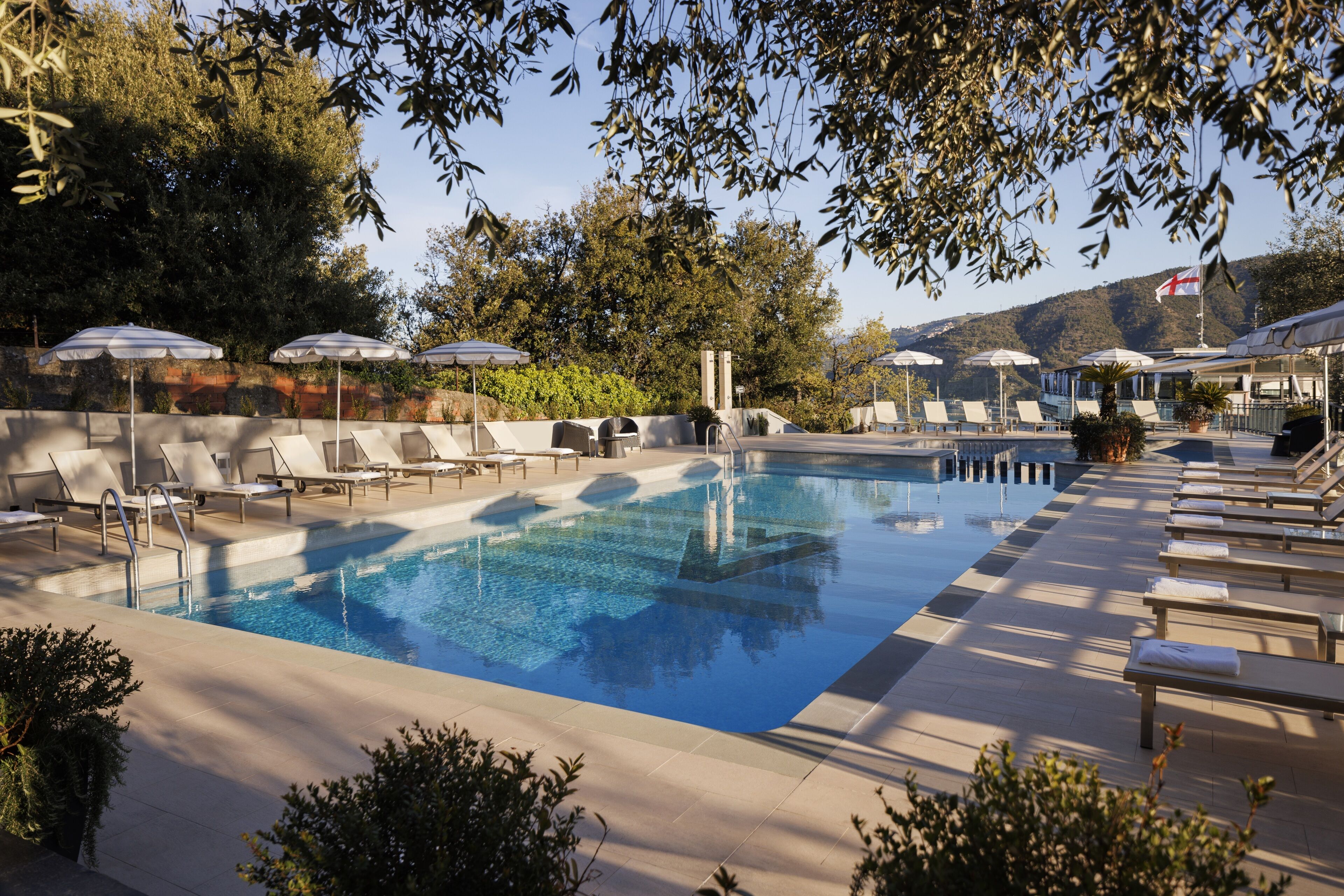 A swimming pool with lounge chairs and umbrellas, surrounded by trees and a mountain in the background.