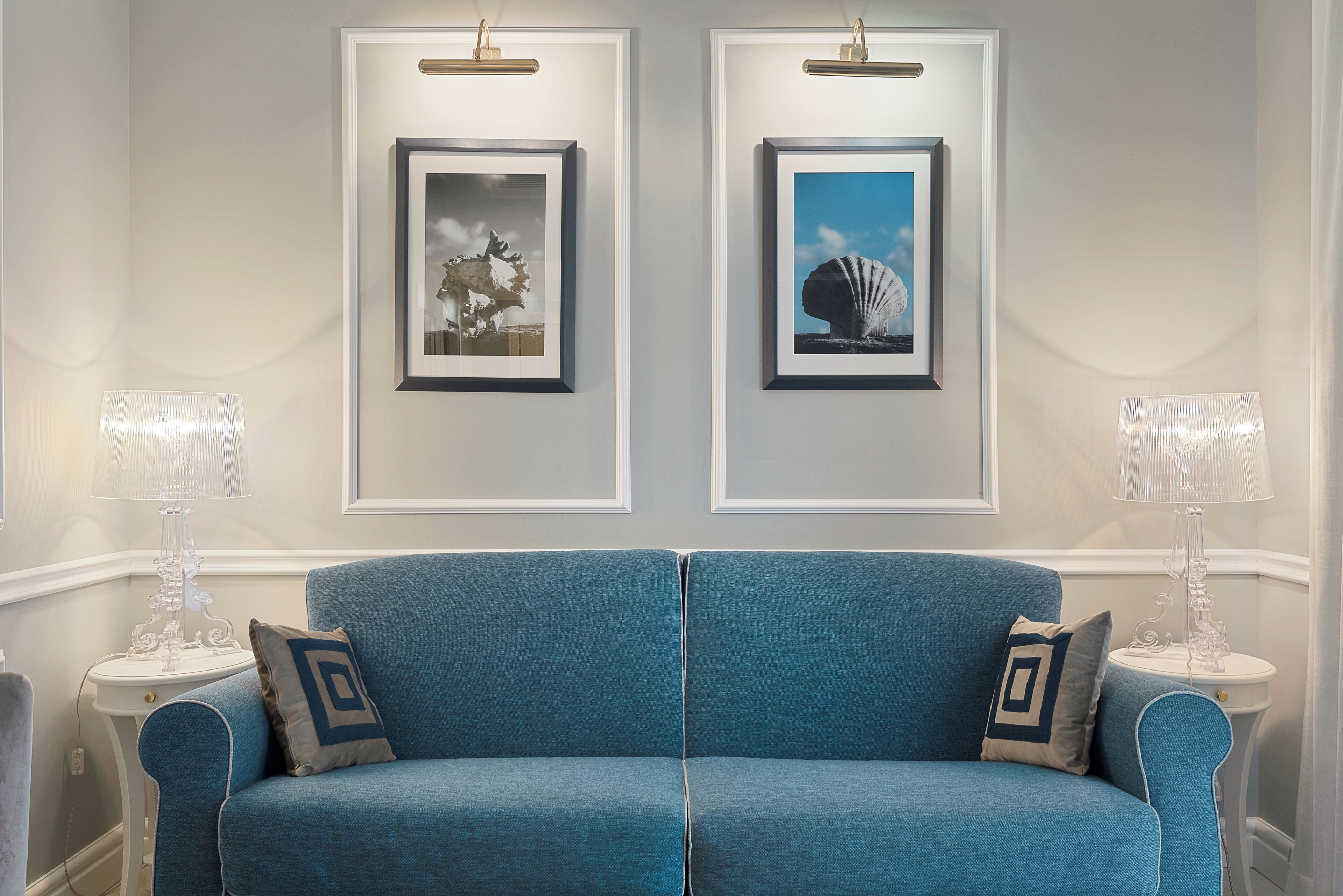 Un sofá azul con dos almohadas, flanqueado por dos mesitas blancas con lámparas, frente a una pared con dos cuadros enmarcados.