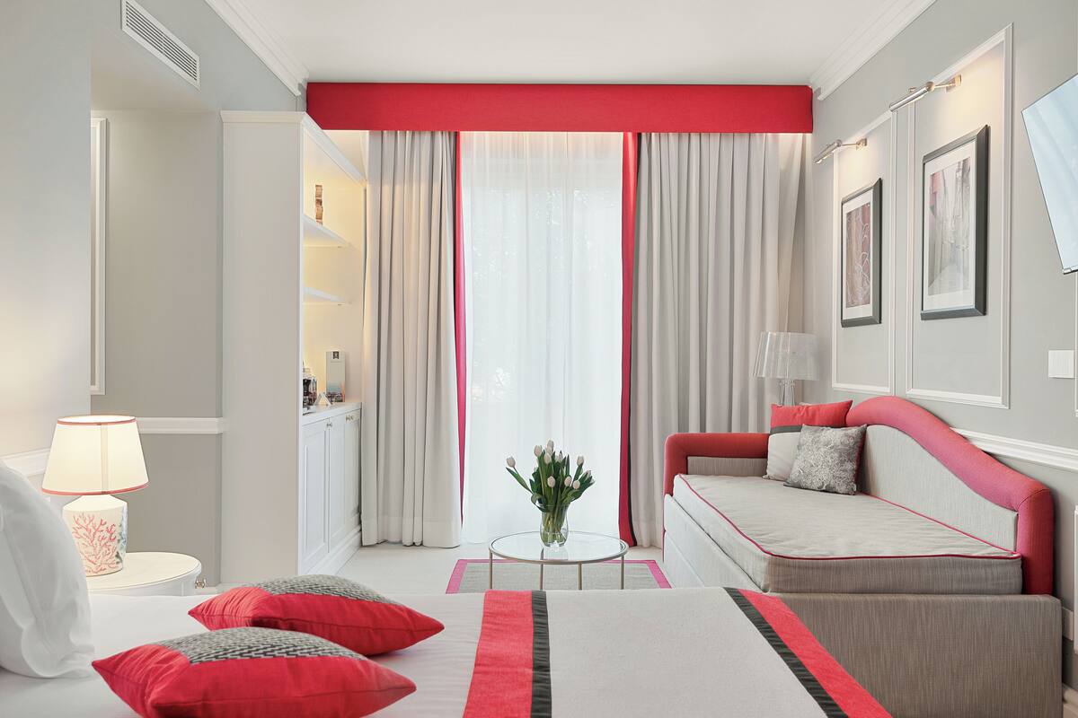 A modern hotel room with a bed, a sofa, a small table with a vase, and a view of the outdoors through large windows.