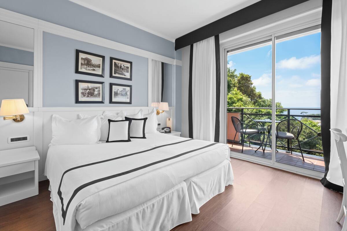 A hotel room with a large bed, a balcony with outdoor seating, and framed pictures on the wall.