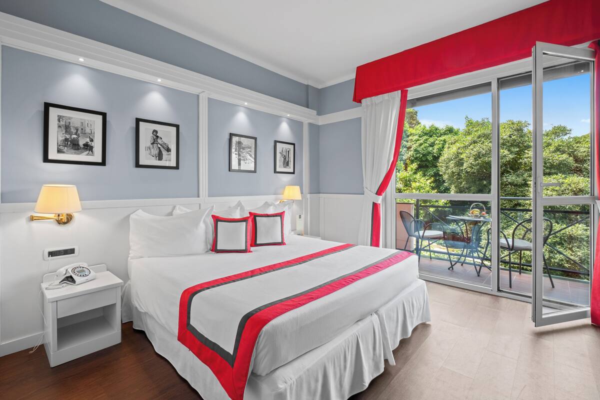 A hotel room with a large bed, two bedside lamps, a nightstand, and a balcony with a table and chairs.