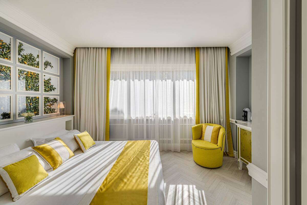 A hotel room with a bed, a yellow armchair, a window with curtains, and a bedside table.