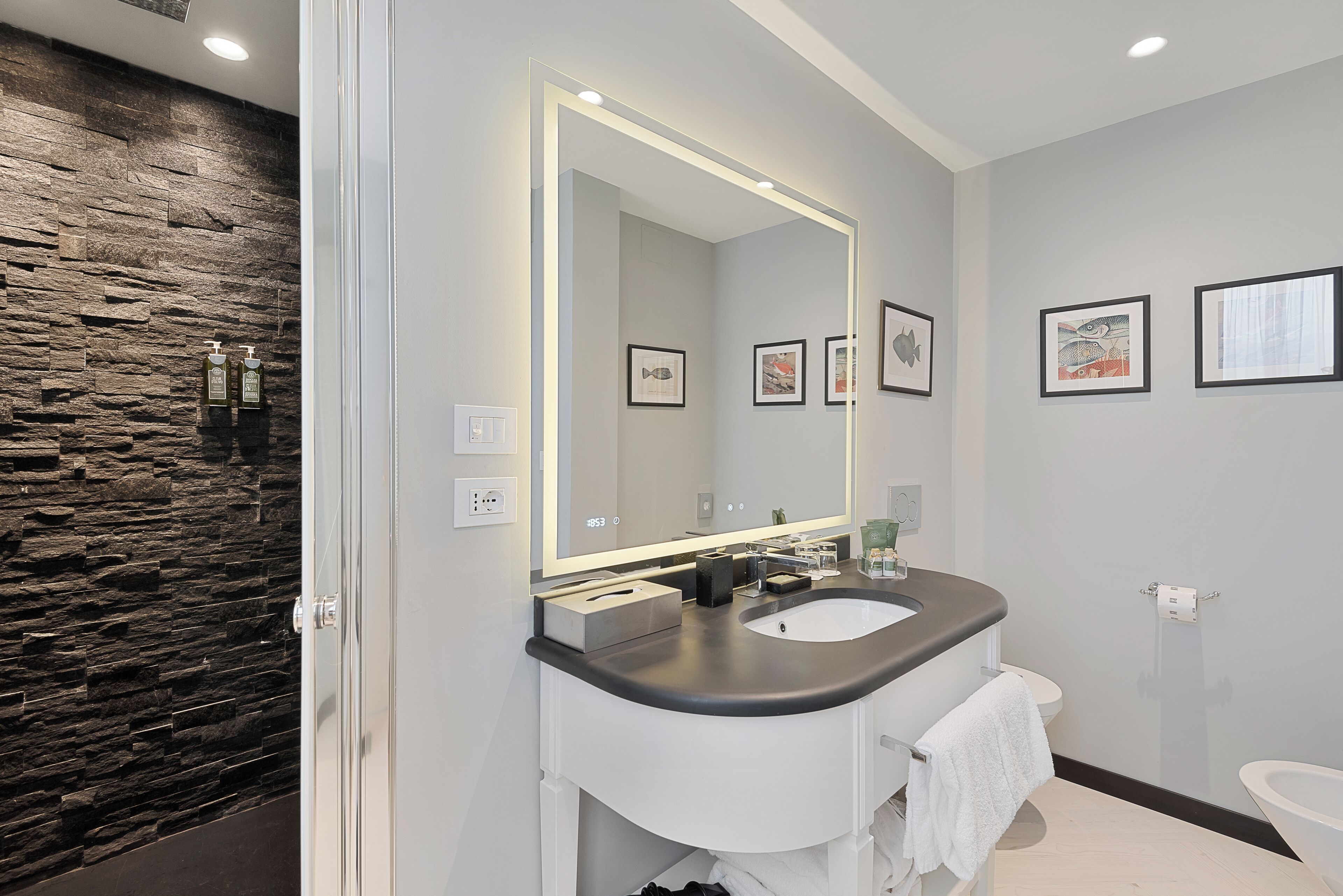 A modern bathroom with a stone wall, a large mirror, a white sink, and framed artwork on the wall.
