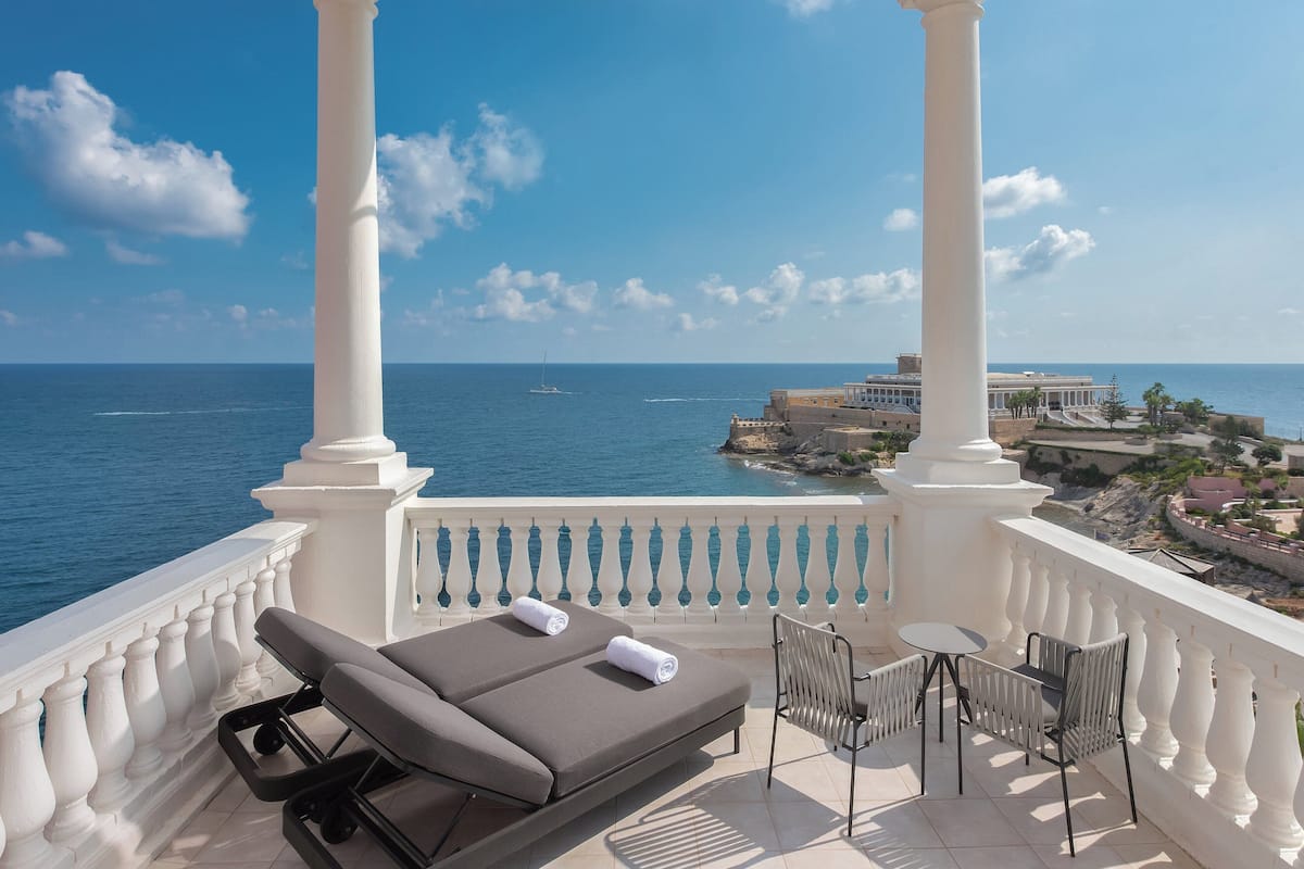 A balcony with a view of the sea, a lounge chair, a table, and chairs.