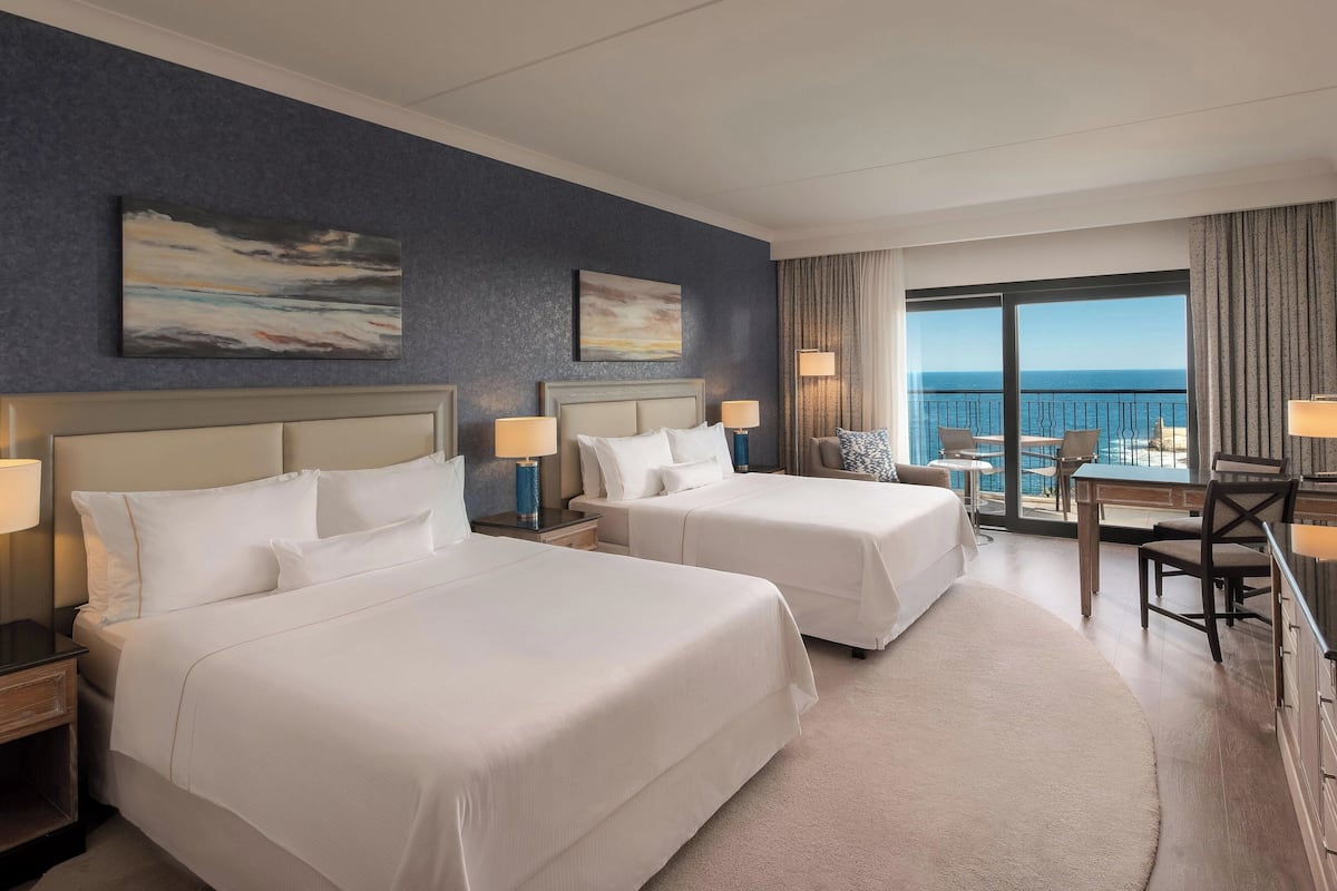 A hotel room with two beds, a desk, and a balcony with a view of the ocean.