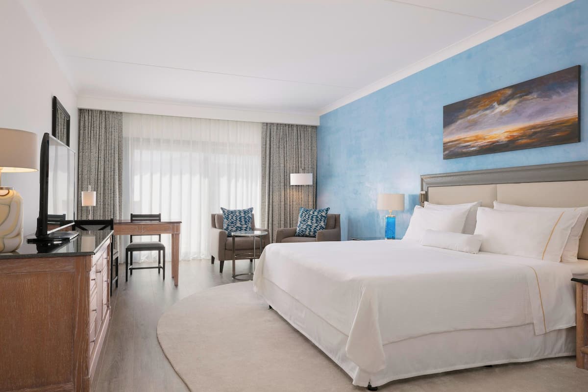 A hotel room with a large bed, a desk, two chairs, a TV, and a painting on the wall.