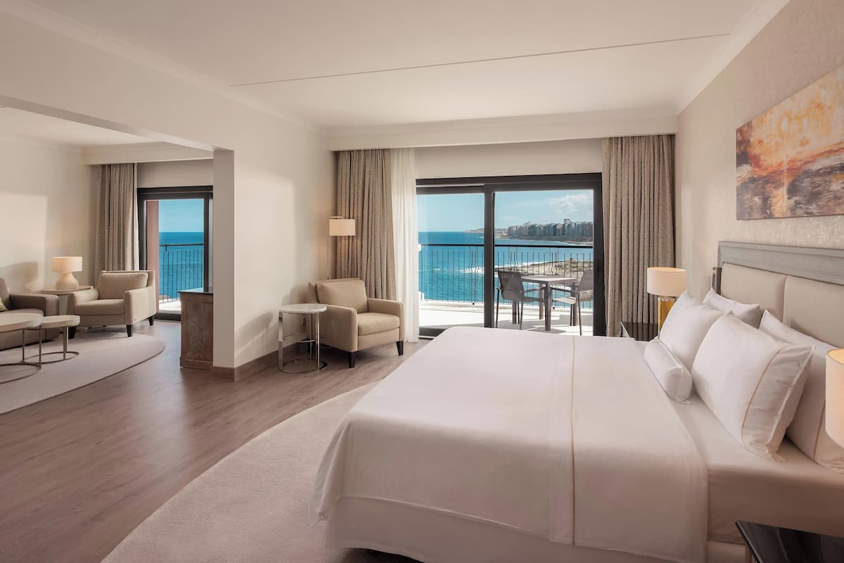 A hotel room with a large bed, two armchairs, a small table, and a balcony with a view of the sea.