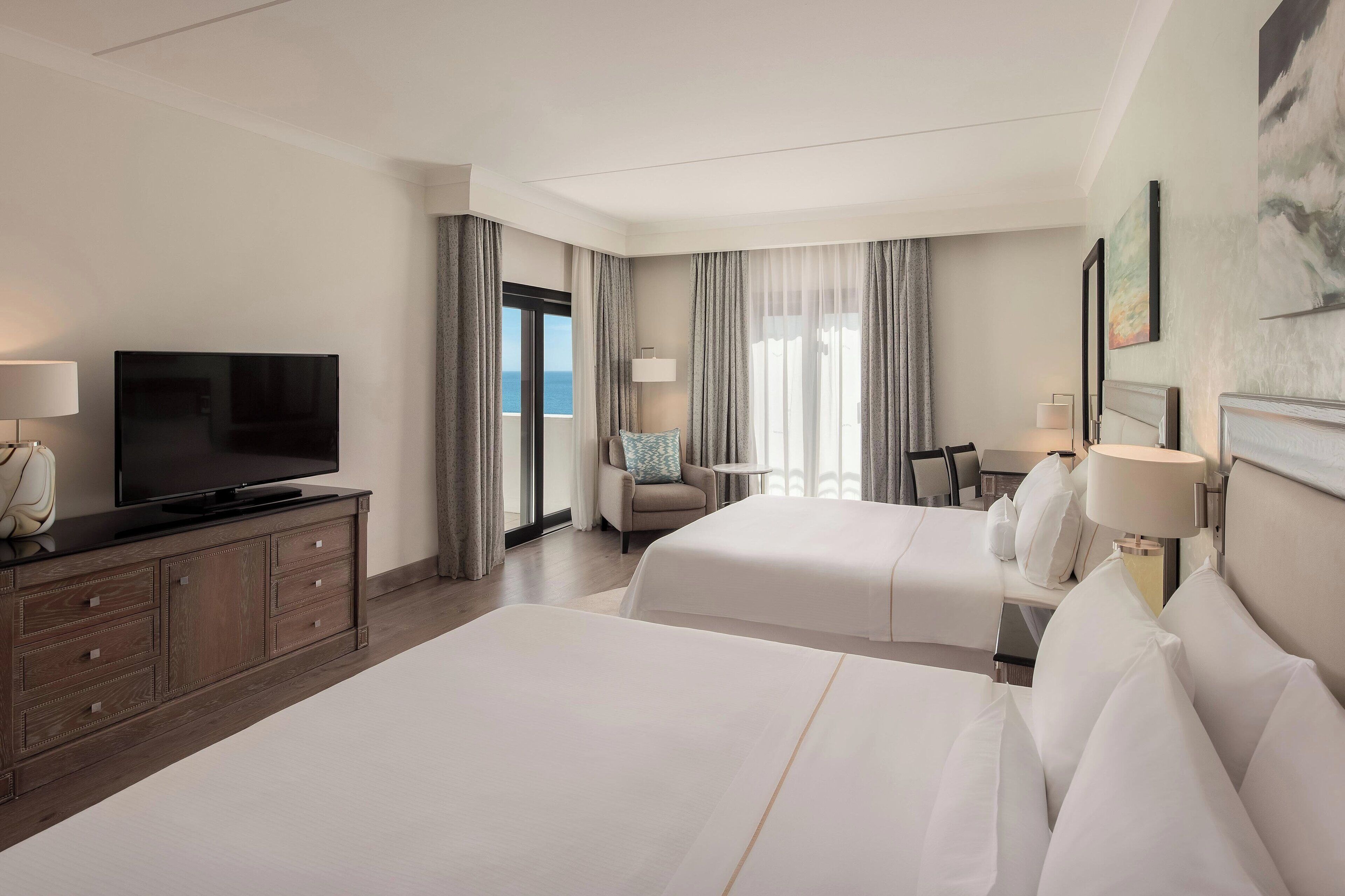 A hotel room with two beds, a TV, a desk, and a view of the ocean.