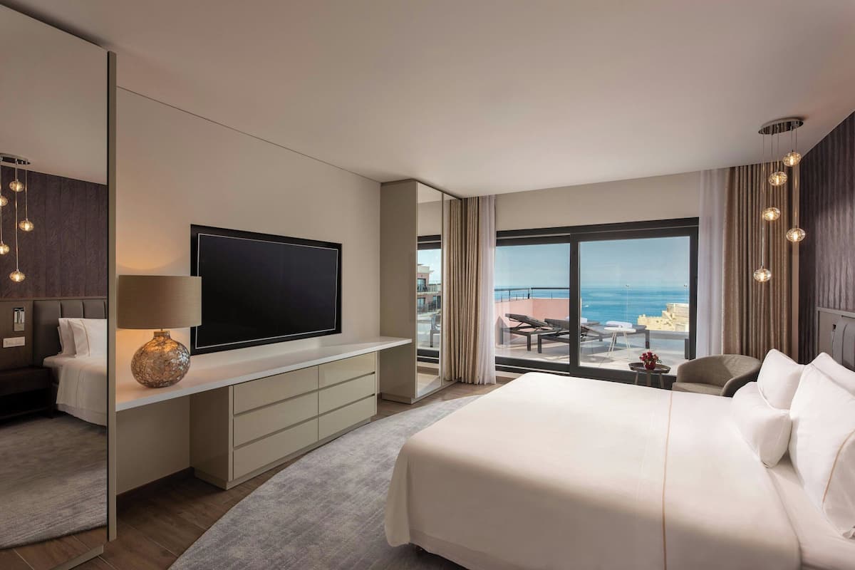 A modern hotel room with a large bed, a flat-screen TV, a desk with a lamp, and a balcony with a view of the sea.