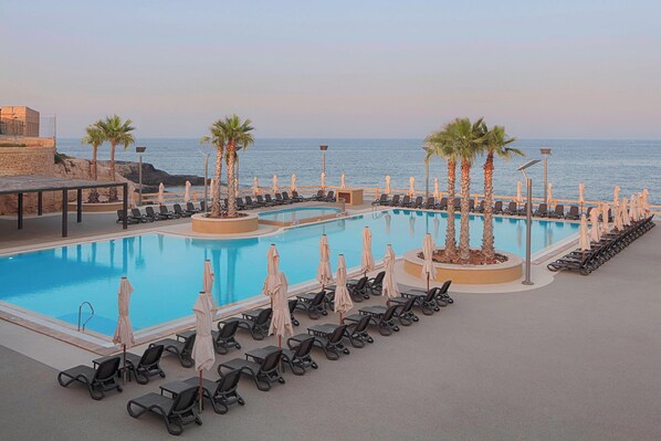 A pool area with multiple lounge chairs and umbrellas, a clear view of the sea, and a stone wall on one side.