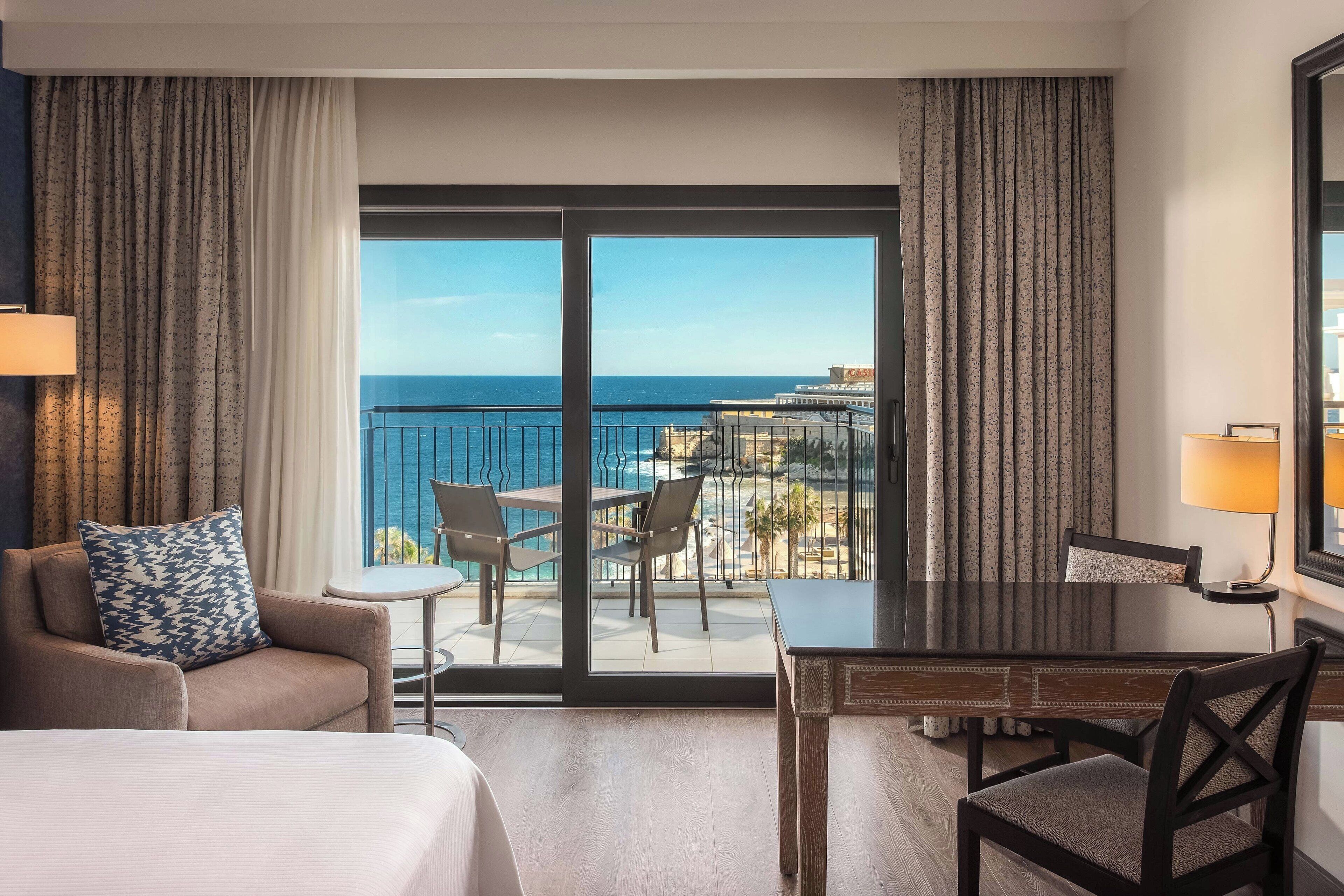 Une chambre d’hôtel comprenant un lit, un bureau, une chaise, une petite table et un balcon avec vue sur l’océan.