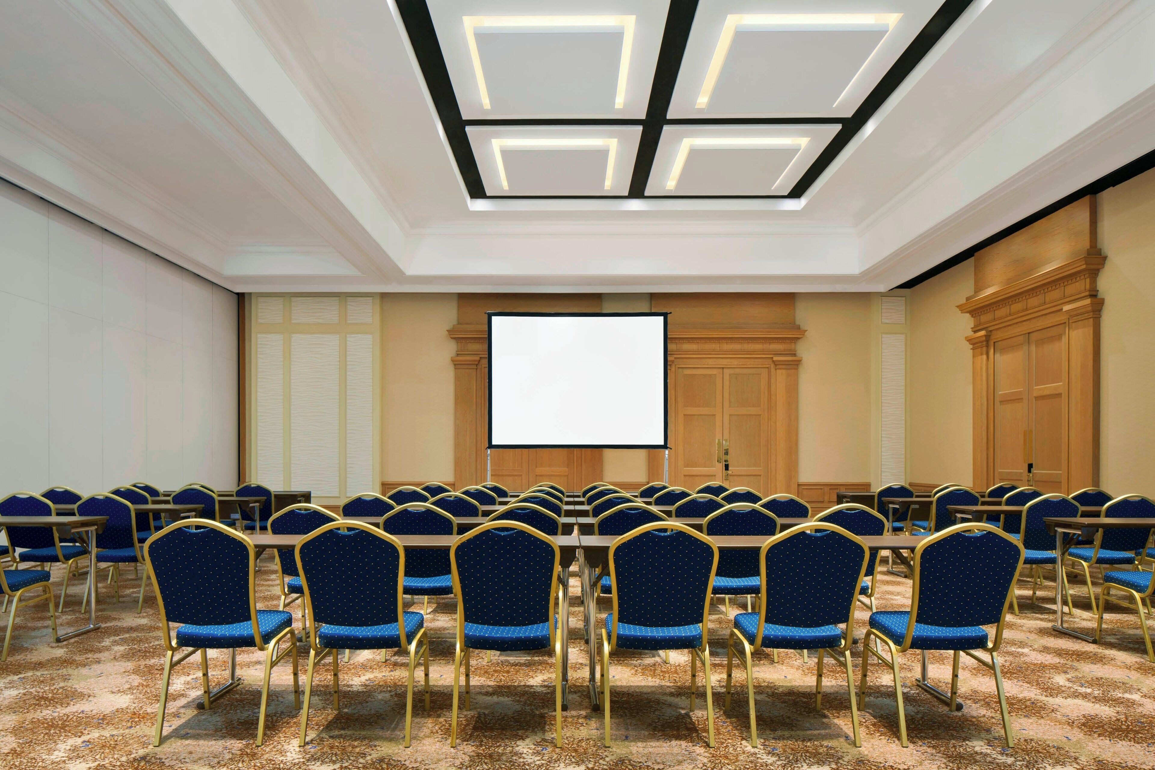 A conference room with a large screen, rows of blue chairs, and a carpeted floor.