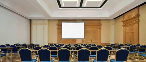 A conference room with a large screen, rows of blue chairs, and a carpeted floor.