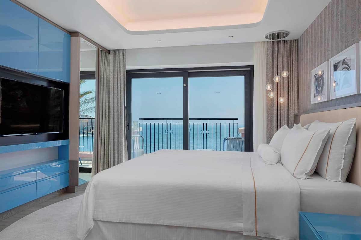 A modern hotel room with a large bed, a flat-screen TV, and a balcony with a view of the sea.