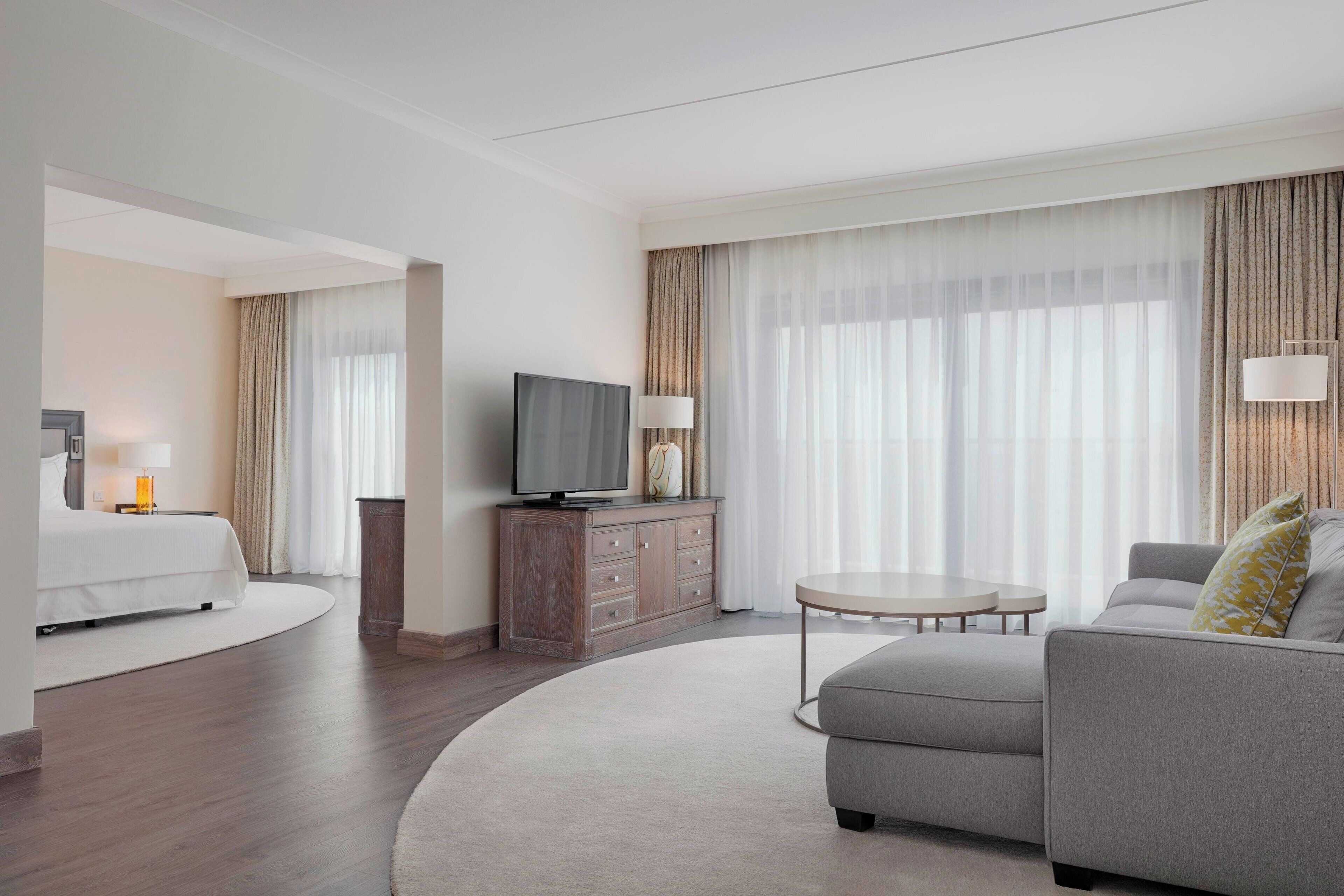 Een moderne hotelkamer met een bed, een bank, een televisie op een houten kast, en een groot raam met dunne gordijnen.