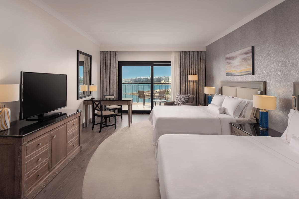 A hotel room with two beds, a TV, a desk, and a view of the city.