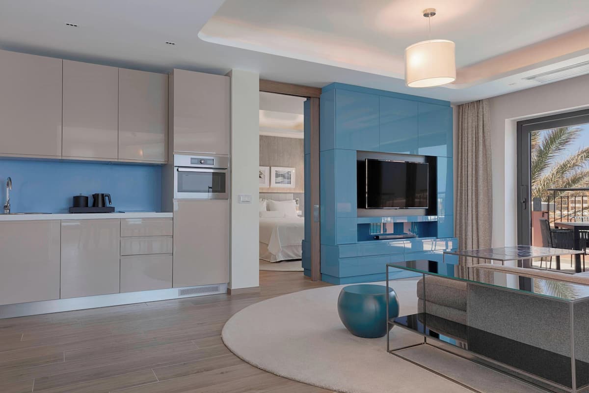 A modern hotel room with a kitchenette, a TV, and a glass dining table.