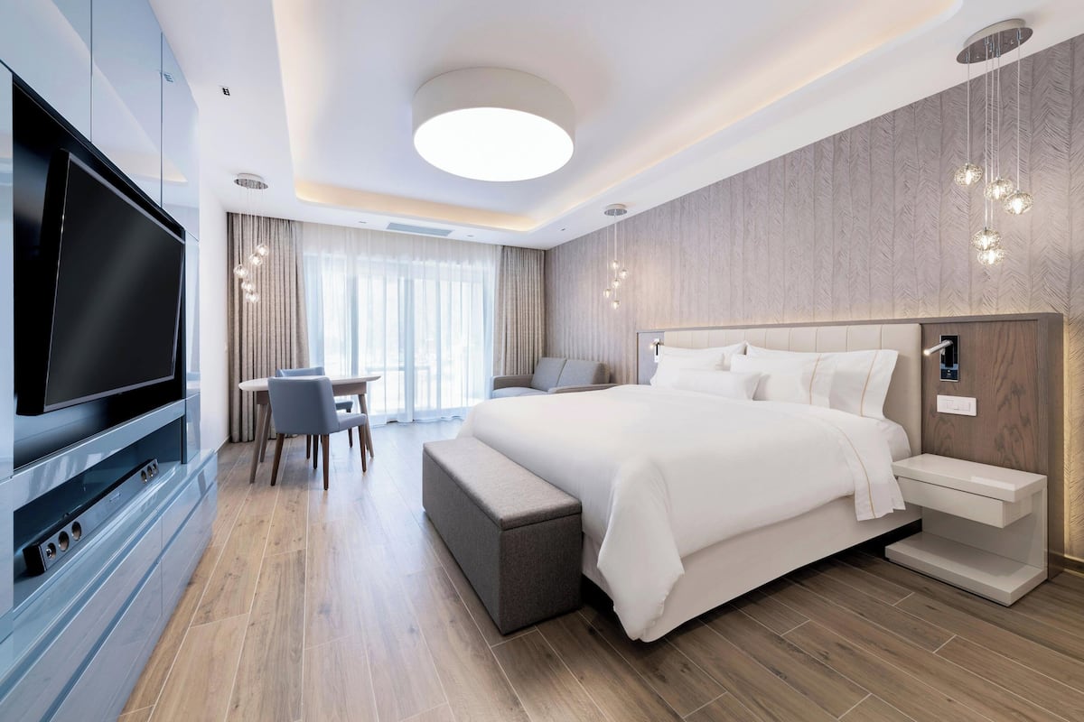 A modern hotel room with a large bed, a flat-screen TV, a seating area with a table and chairs, and a large window with curtains.