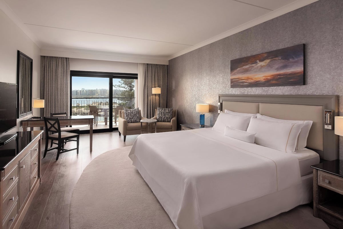 A hotel room with a large bed, a desk with a chair, a small table with chairs, and a view of the city through large windows.