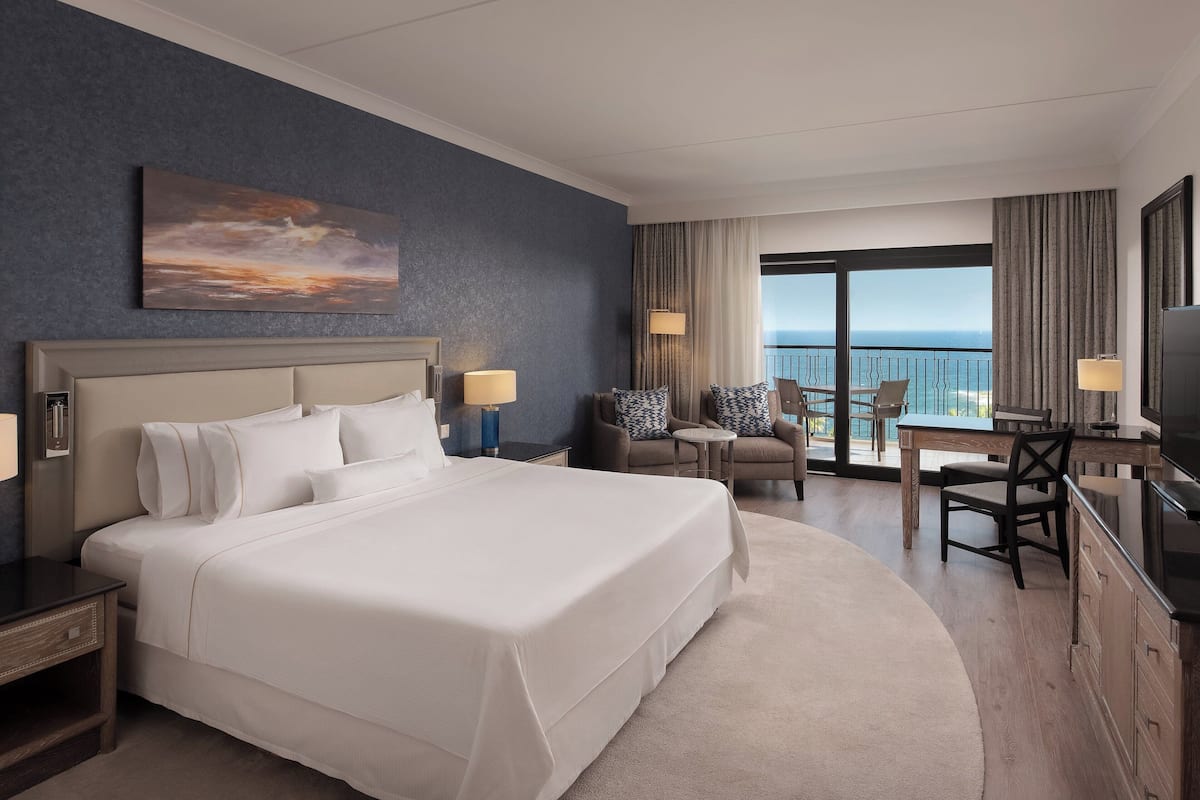 A hotel room with a large bed, a desk with a chair, a television, and a balcony with a view of the ocean.
